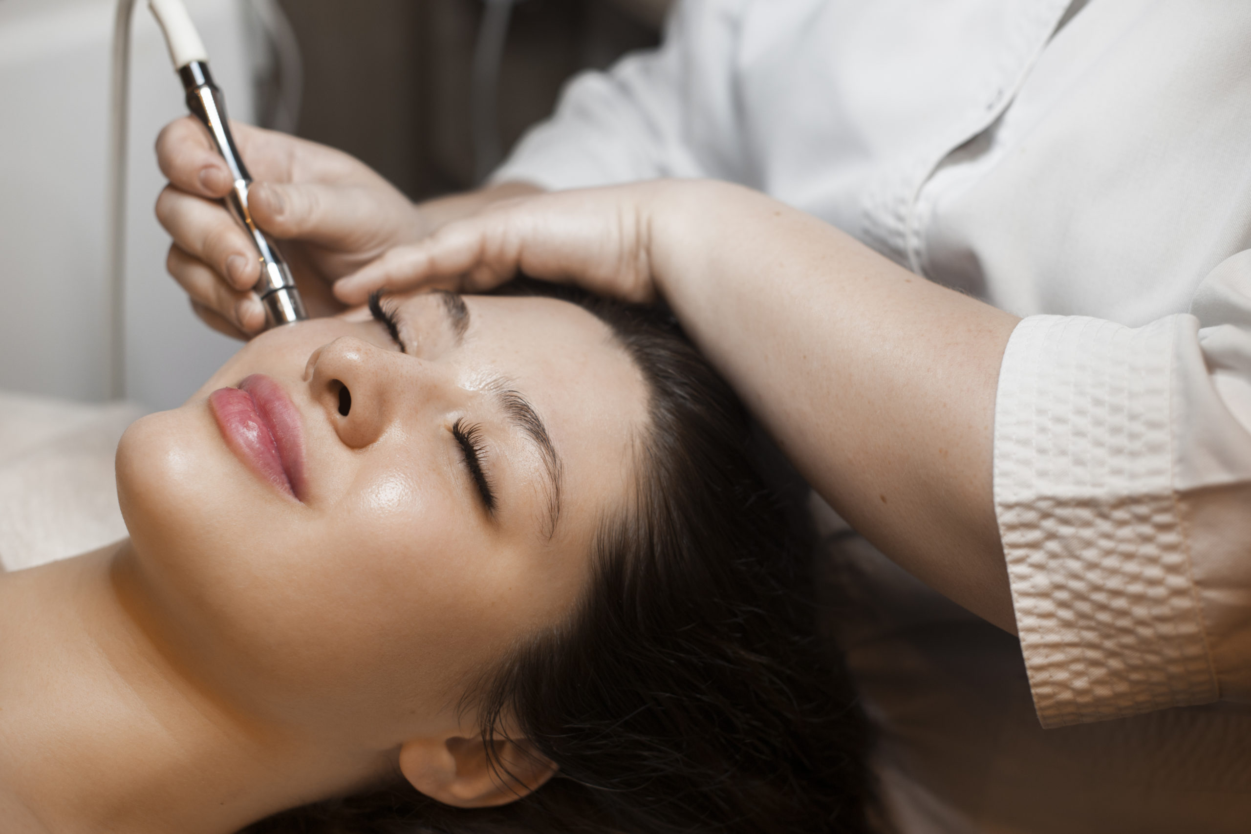 Close up side view portrait of a cute caucasian woman doing noninvasive microdermabrasion on her face with a dermapen by a cosmetician.